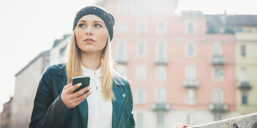 A young blonde woman wearing a hat and a leather jacket stands in a city holding a smartphone in her hand. A blurred building can be seen in the background, and the mobile-pocket logo is located in the bottom right corner.