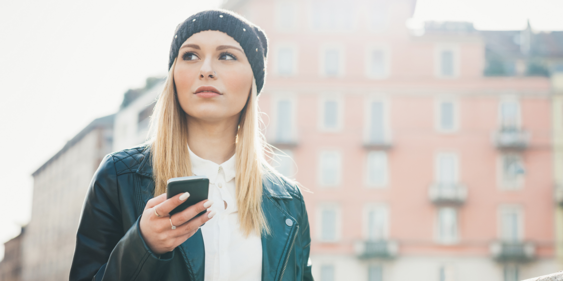 Eine junge blonde Frau mit Mütze und Lederjacke steht in einer Stadt und hält ein Smartphone in der Hand. Im Hintergrund ist ein unscharfes Gebäude zu sehen, unten rechts befindet sich das mobile-pocket Logo.