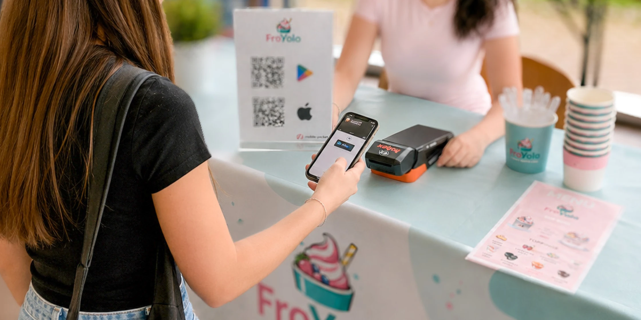 The picture shows the FroYolo stall set up as part of a school project: a girl is making a simple payment using her smartphone at the hobex payment terminal and the mobile-pocket app, which is designed for digital customer loyalty.
