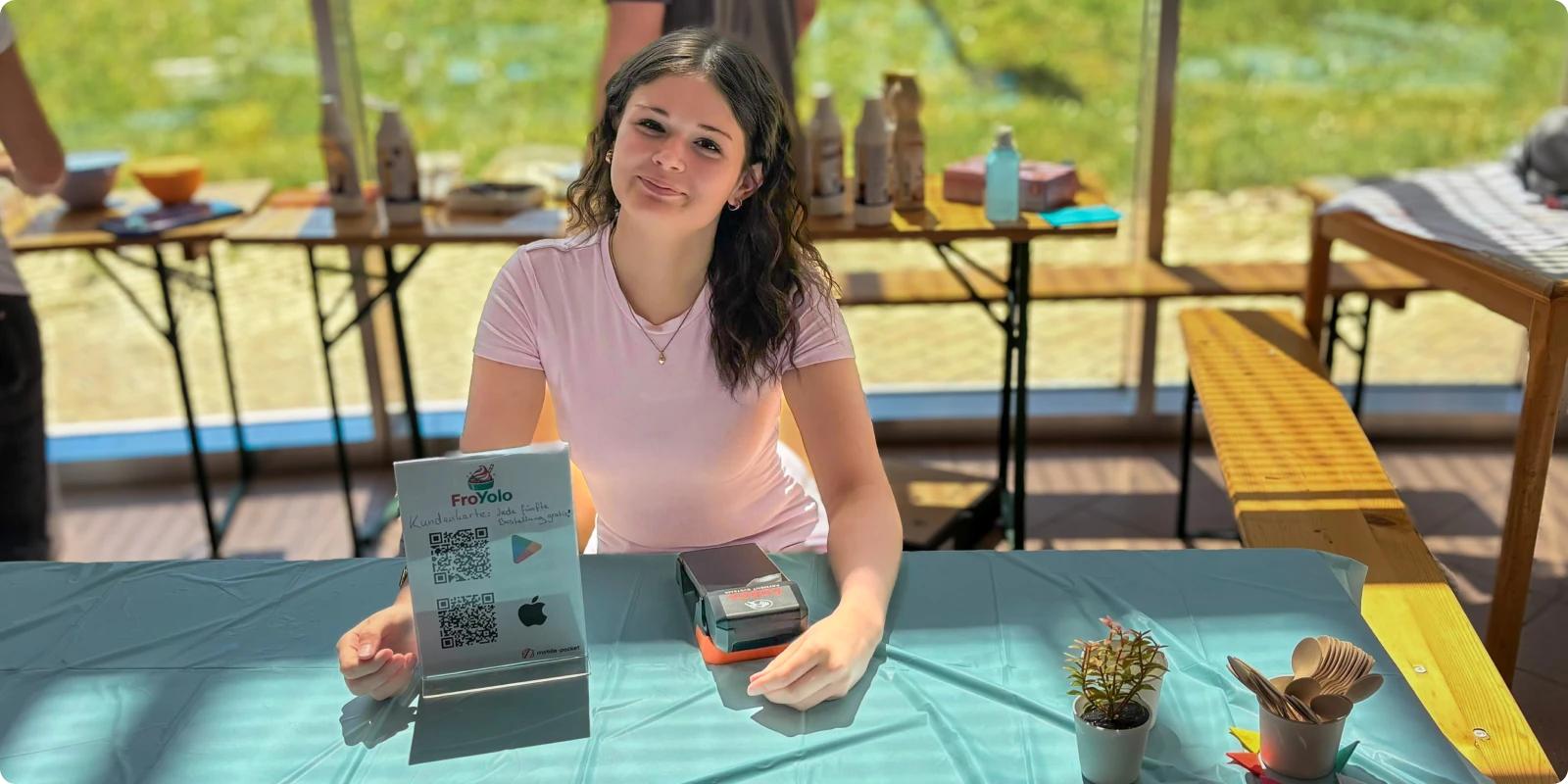 The photo shows the sales stand of the school project training firm FroYolo: co-founder Lena with the hobex sales terminal and a display stand featuring QR codes for quick access to the mobile-pocket customer loyalty app. A digital sales process and loyalty scheme made simple.