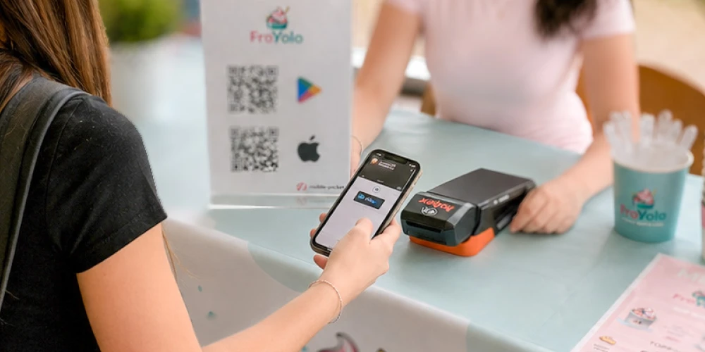 Das Bild zeigt den FroYolo Verkaufsstand eines Schulprojekts: Ein Mädchen tätigt mit dem Smartphone einen einfachen Bezahlvorgang am Bezahlterminal hobex und mobile-pocket, der App für digitale Kundenbindung. 