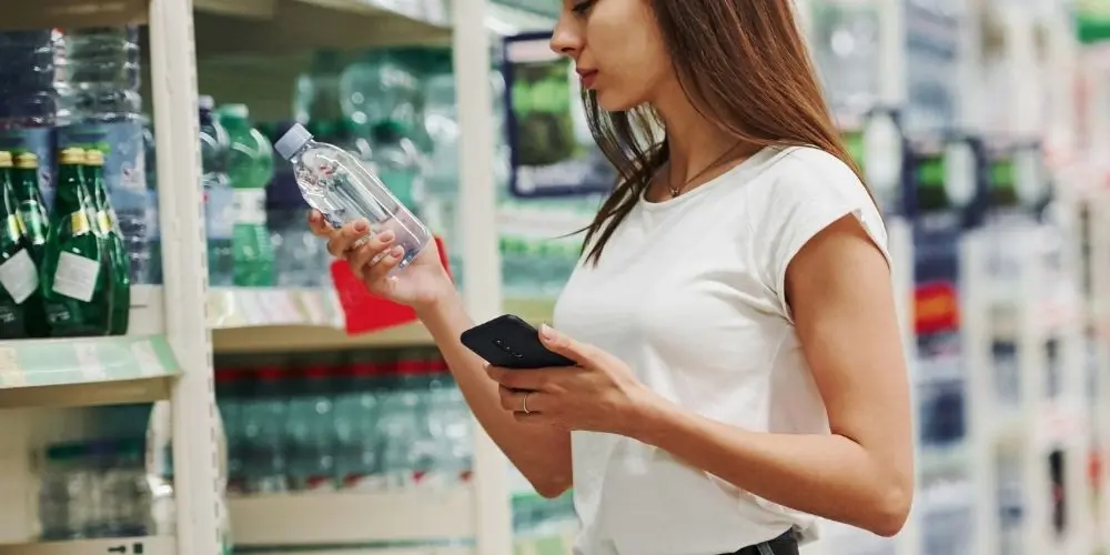A young woman is standing in front of the drinks shelf in the supermarket, holding a bottle of drink in one hand and her smartphone in the other. An example for promoting a loyalty app through specific mobile touchpoints.